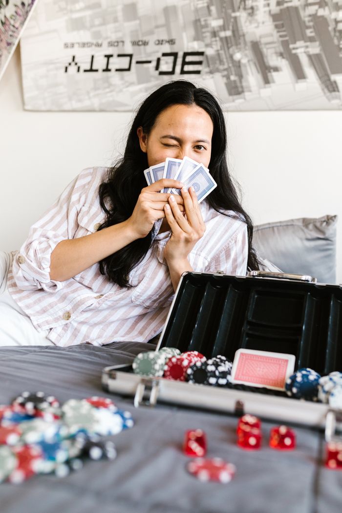 A woman sits on a bed holding playing cards, with poker chips and dice around her.